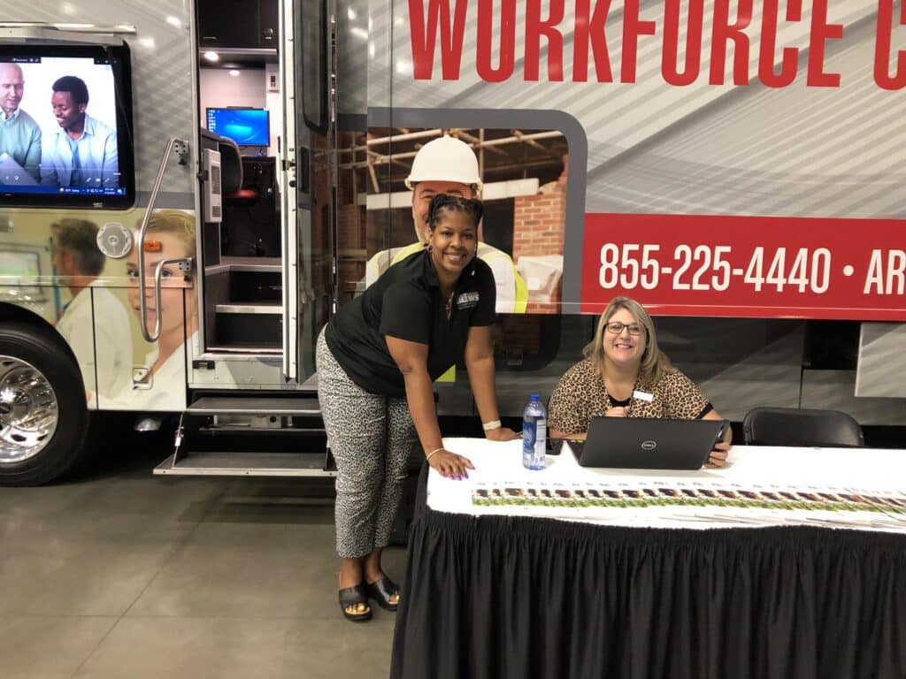 A female DWS employee sitting at a table and another female DWS employee standing next to her along the outside of a large mobile workforce center vehicle parked inside an arena